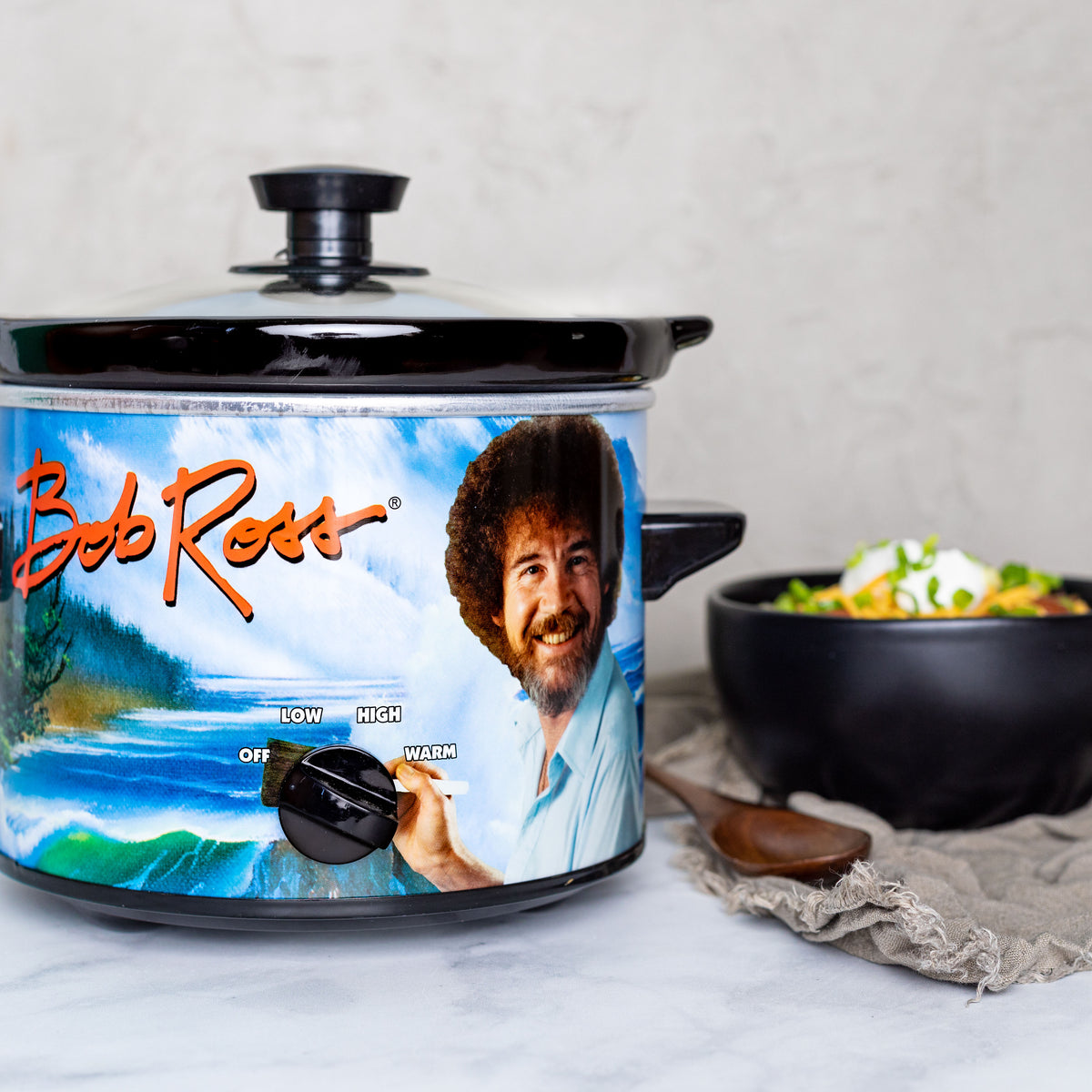 Bob Ross slow cooker in focus, bowl of chili in the background